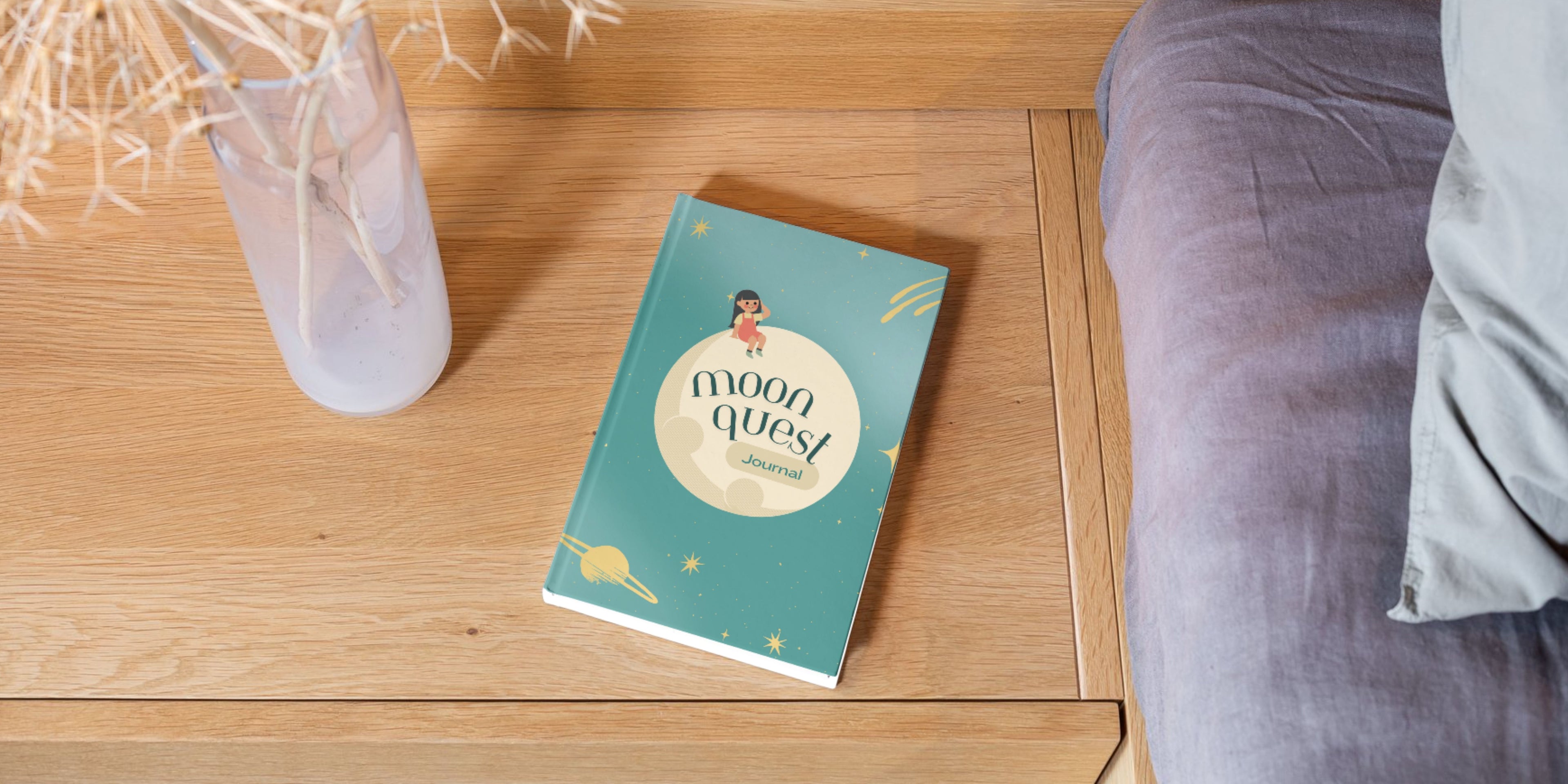Journal titled 'Moon Quest' on a wooden surface with a vase and the edge of a mattress and pillow in the background